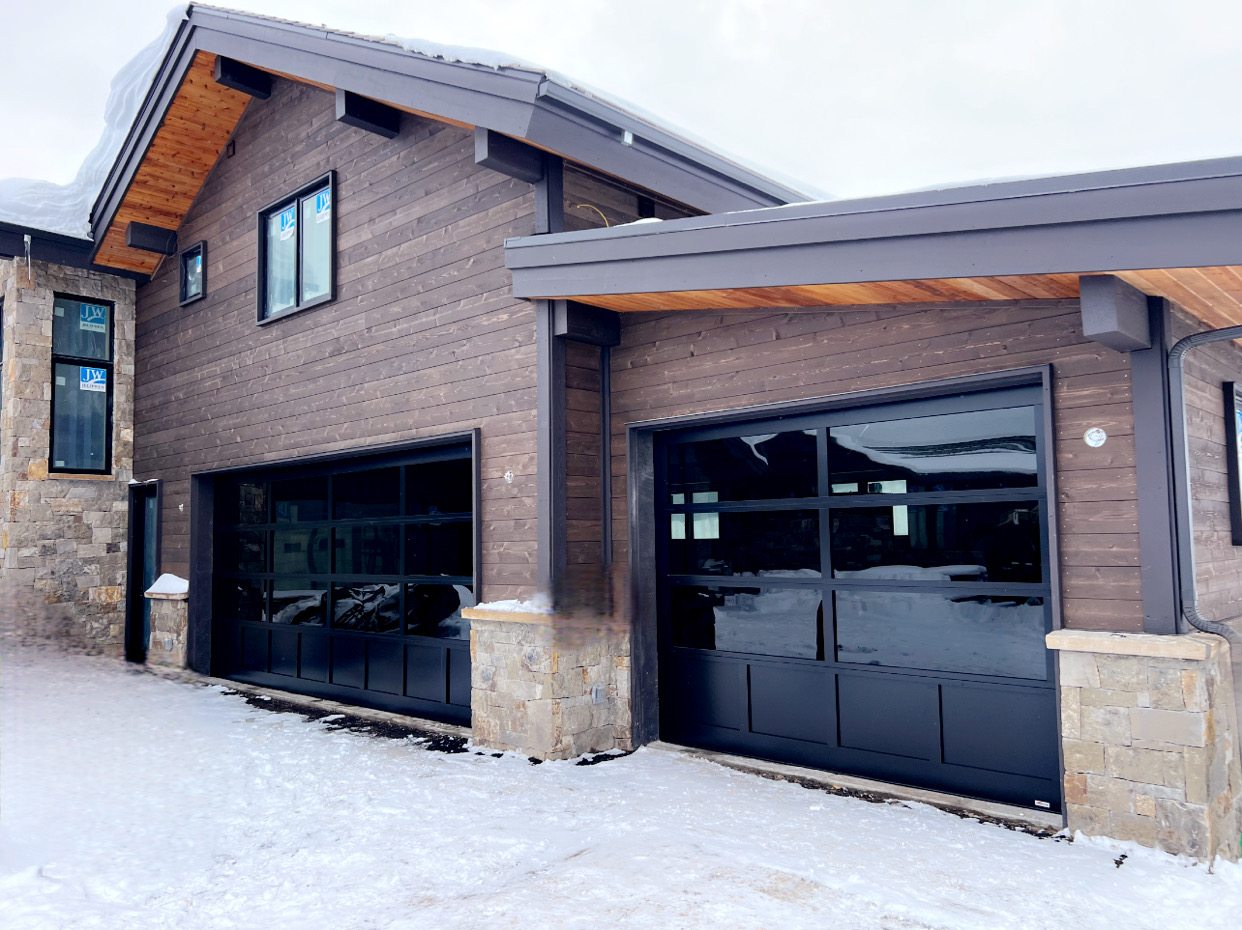 Two black full-view glass garage doors installed on a wood-sided mountain home in the snow